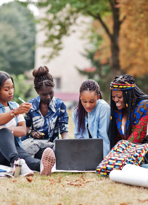 Group of five african college students spending time together on campus at university yard. Black afro friends sitting on grass and studying with laptops.